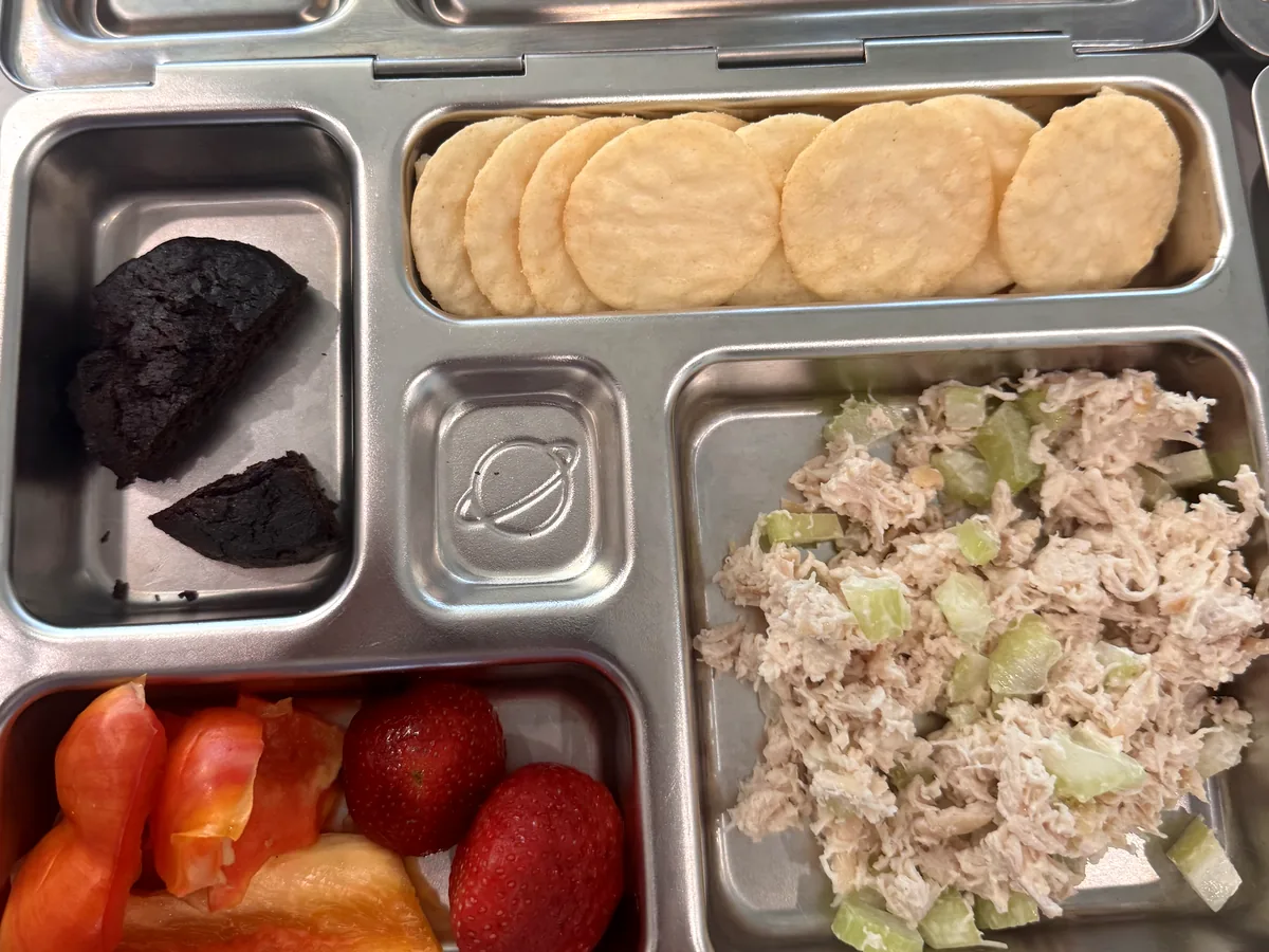 A steel bento box with chicken salad, round crackers, brownie pieces, strawberries and red pepper