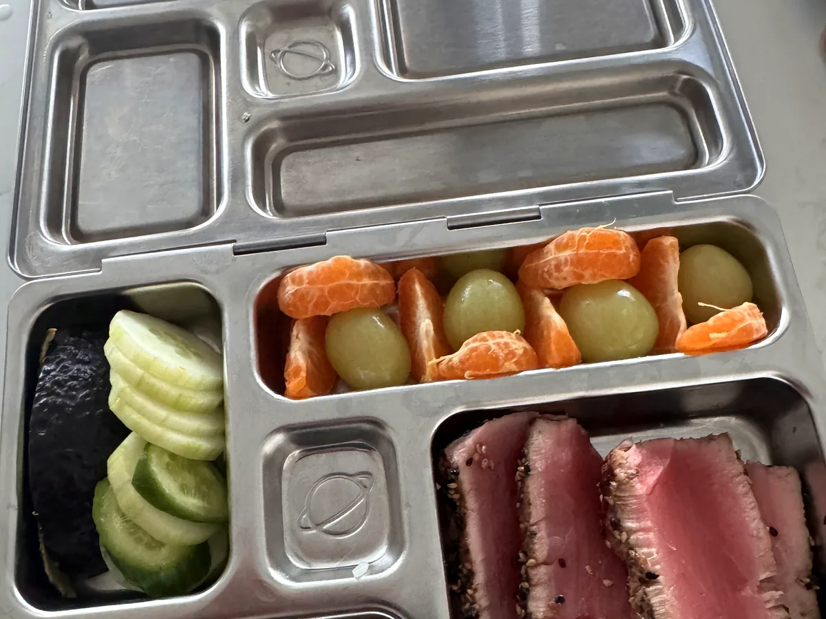 A steel bento box with sliced seared tuna, rice, cucumber slices, clementine segments and grapes
