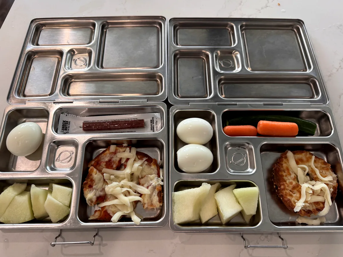 Two steel bento boxes with breaded chicken, boiled eggs, melon and carrots