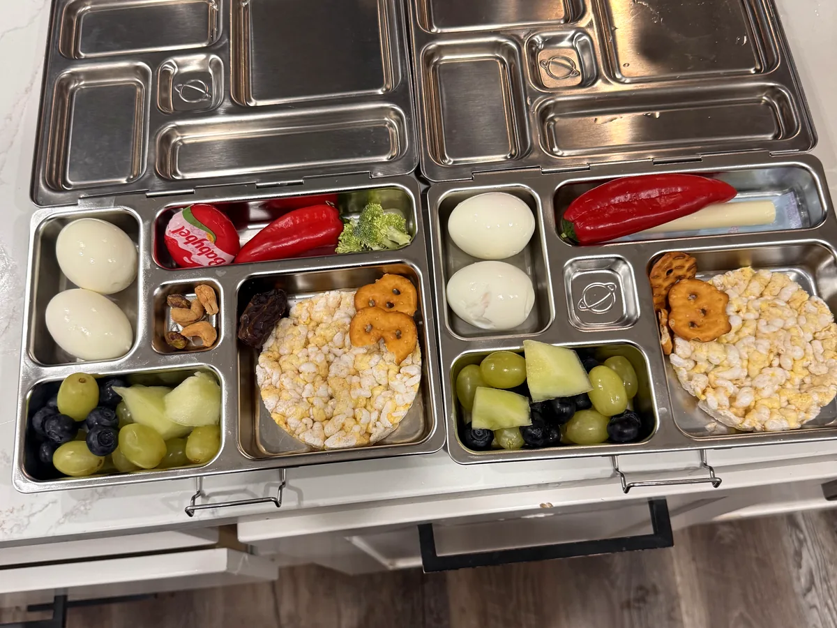 Two steel bento boxes with boiled eggs, popcorn, melon, peppers and a meat stick