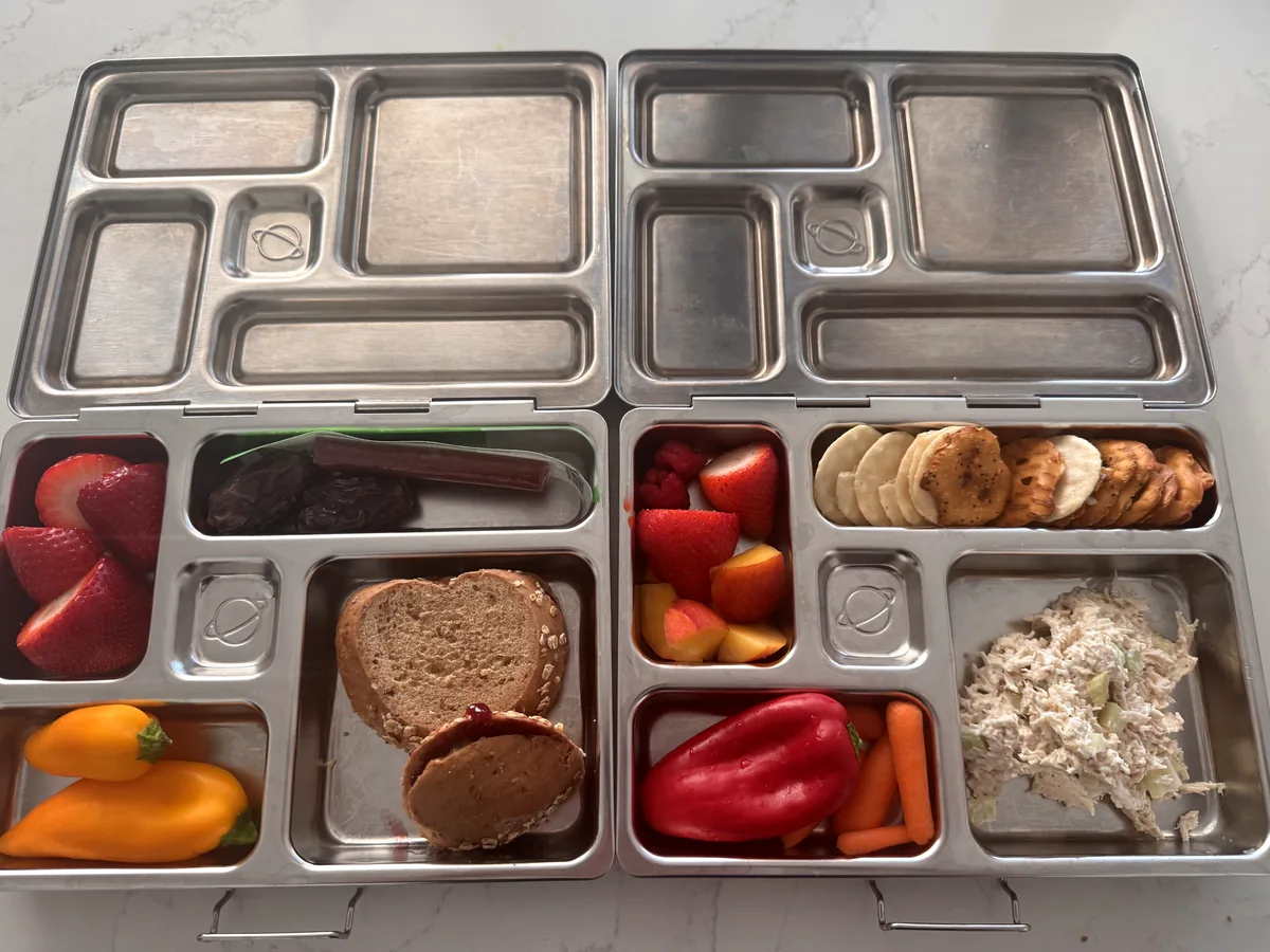 Two steel bento boxes side by side, one with a heart shaped sandwich and strawberries, one with chicken salad, peppers and banana chips