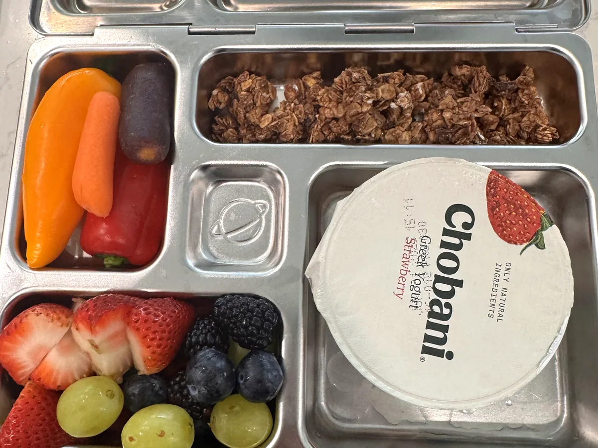 A steel bento box with a yogurt cup, granola, berries and grapes, carrots and peppers